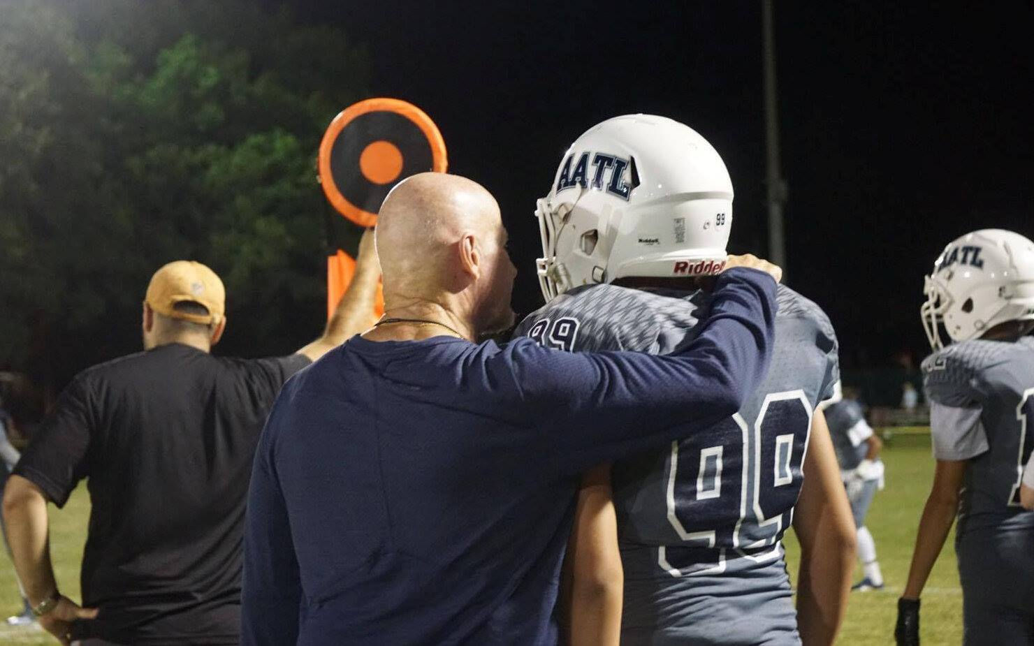A coach with his arm around a young player’s shoulder pads on the sideline at night.